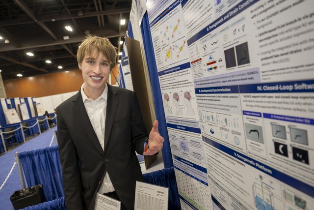 Official ISEF photo of Evan in a dark suit standing beside his SILENCE project poster board in the convention hall, smiling and giving a thumbs-up