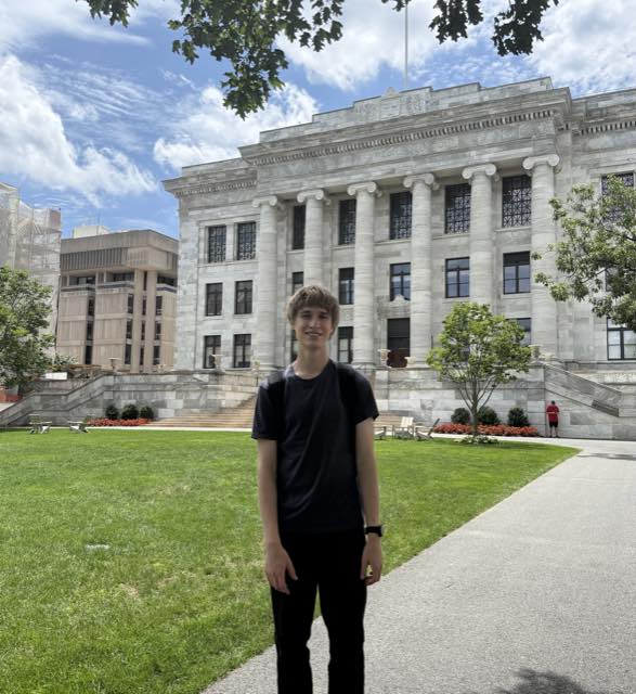 Evan on the lawn in front of a white marble neoclassical hall with tall Ionic columns on a bright summer day
