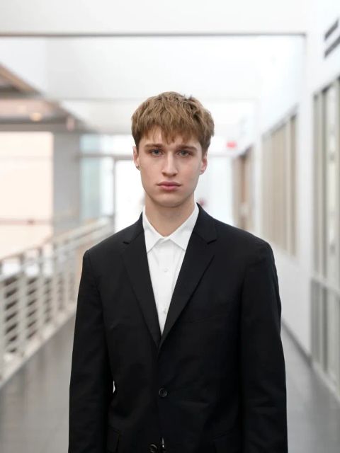 Portrait of Evan in a black suit and white open-collar shirt, standing in a bright glass-walled walkway