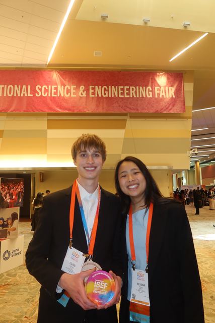 Evan, wearing a medal and lanyard badge, with a female finalist in front of a red 'International Science & Engineering Fair' banner; he holds a rainbow ISEF award disc