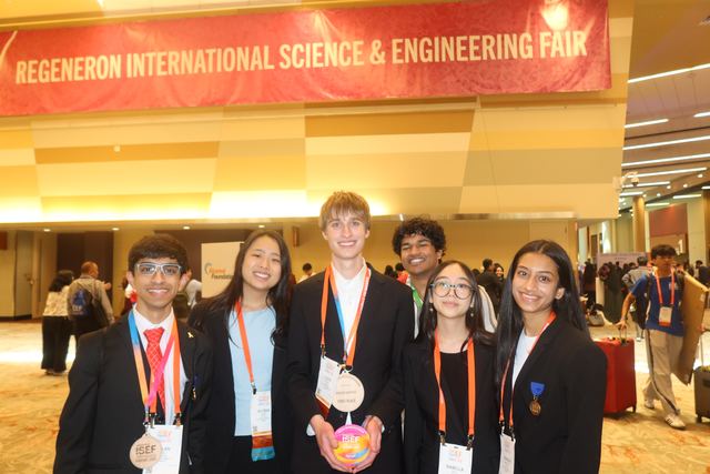 Group of six finalists posed beneath the full 'Regeneron International Science & Engineering Fair' banner, Evan center holding the rainbow ISEF award disc, one student wearing a medal