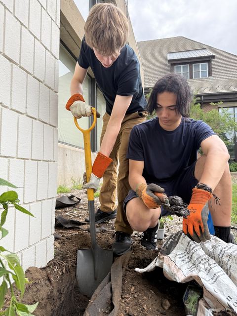 Evan digging with a yellow-handled shovel while a friend in work gloves crouches beside him sorting soil, planting along a white brick building wall