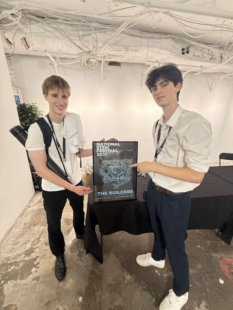 Evan and another young man in white dress shirts holding a framed 'National STEM Festival 2026, The Builders, Washington, DC' poster in a backstage-style room with exposed ceiling conduits