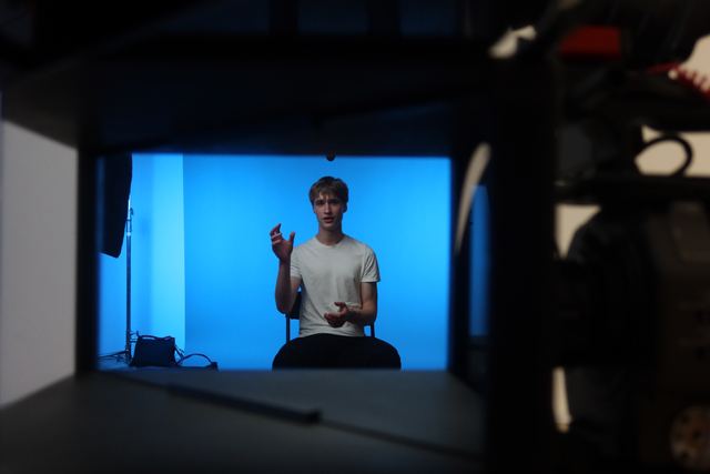Behind-the-scenes shot of Evan seated against a bright blue studio backdrop mid-gesture during an on-camera interview, framed through the silhouetted camera rig and lighting equipment