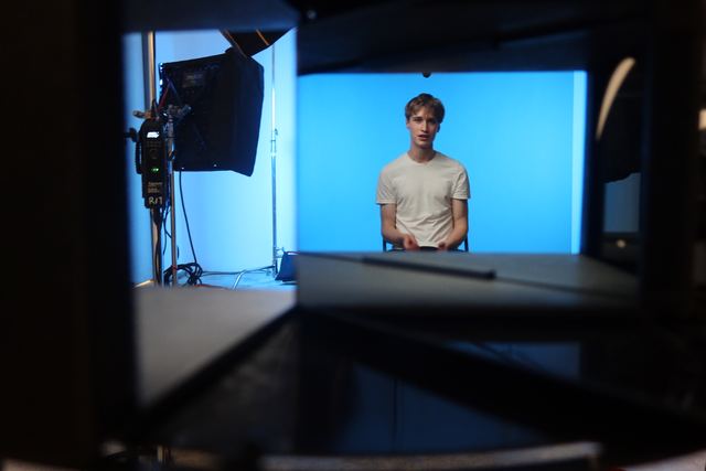 Behind-the-scenes view through camera rigging of Evan seated in front of a bright blue backdrop in a professional video studio with softbox lighting