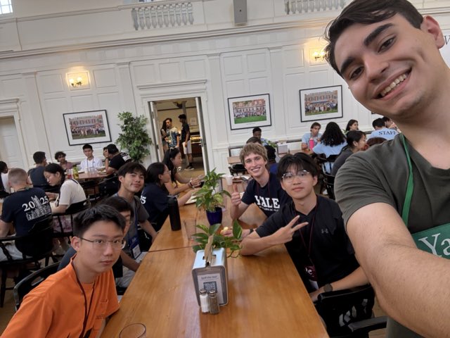 Group selfie at a long communal table in a bright, wood-paneled Yale dining hall during a summer program