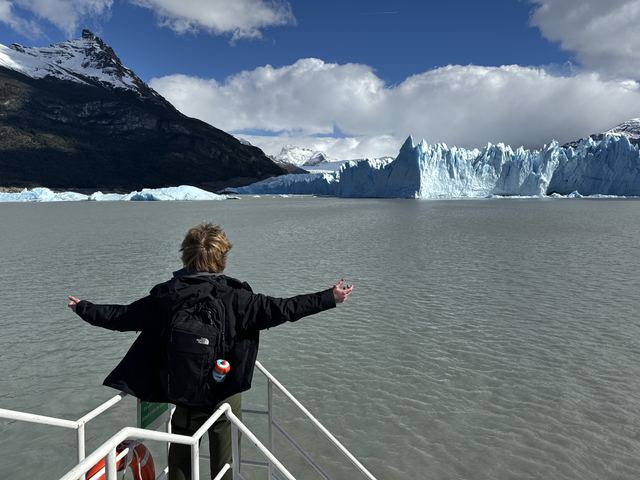 Evan Morris standing at the rail of a boat with arms outstretched, facing the Perito Moreno Glacier in Patagonia