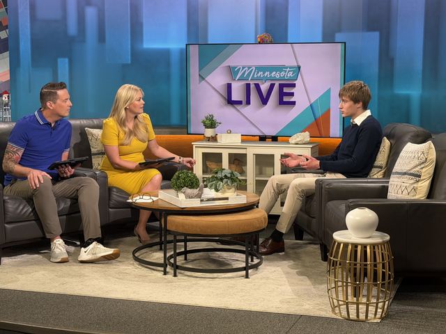 Professional-looking TV studio shot of Evan seated on a couch being interviewed by two hosts on the Minnesota Live set, show logo visible on the screen between them