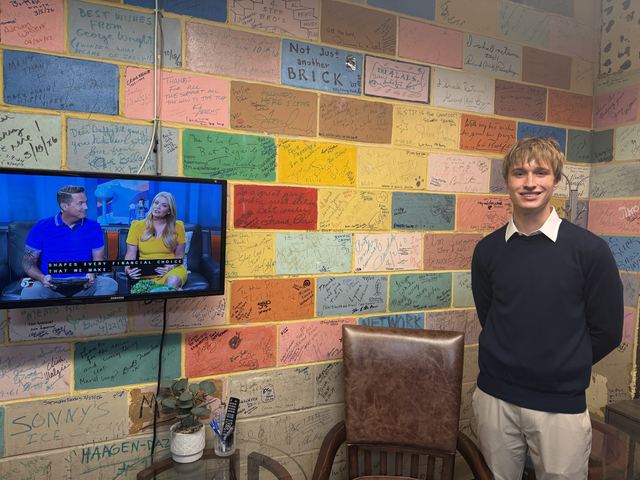 Evan stands smiling beside the station's autographed brick wall while a wall-mounted TV shows the live broadcast with two hosts
