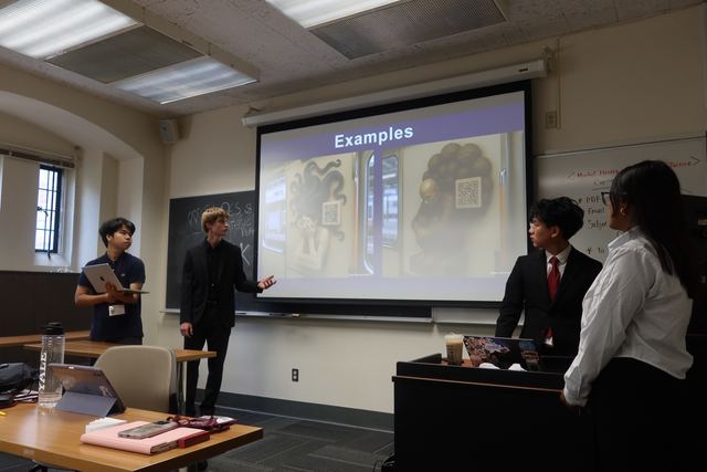 Evan in a dark suit presents an 'Examples' slide to classmates in the same classroom, gesturing at the screen while three teammates look on