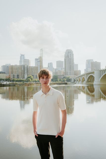 Striking professional portrait standing on a rock at the Mississippi riverfront, cream knit polo, with the full Minneapolis skyline and Third Avenue Bridge mirrored in still water behind him