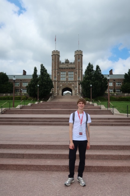 Evan on the wide stone steps below a collegiate Gothic gatehouse tower on a university campus, wearing a visitor lanyard
