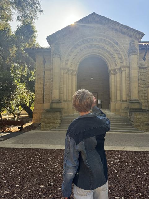 Evan photographed from behind in a denim jacket, facing a sunlit Romanesque sandstone archway building with steps; architecture strongly resembles the Stanford University campus