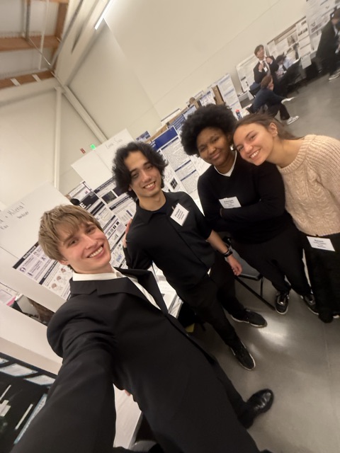 Selfie of Evan in a suit with three other finalists among the poster boards at a science fair