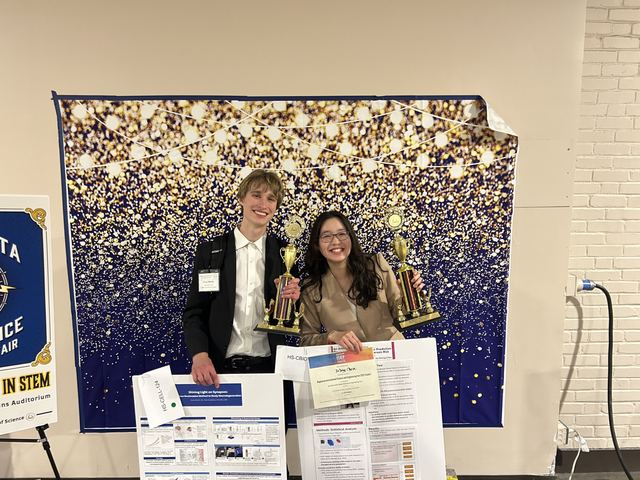 Evan in a suit and a fellow student each holding large gold trophies in front of a navy-and-gold backdrop at the Minnesota State Science & Engineering Fair, with their research posters and certificates in front of them