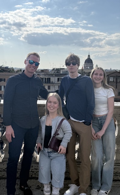 Evan with three others on a sunlit Roman terrace, rooftops and a domed church across the city behind them