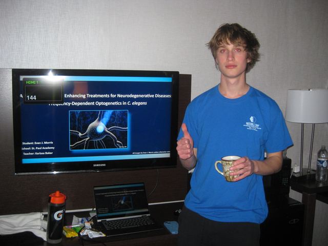 A younger Evan gives a thumbs up beside a hotel-room TV showing his earlier science fair title slide: 'Enhancing Treatments for Neurodegenerative Diseases: Frequency-Dependent Optogenetics in C. elegans', listing his full name, St. Paul Academy, and his teacher's name