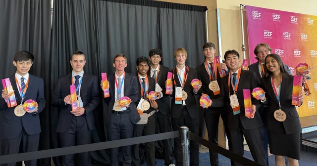 A line of Regeneron ISEF finalists in suits, each holding a rainbow award disc, in front of the ISEF backdrop in the convention hall