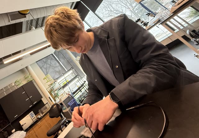 Evan in a dark jacket working over a lab bench, soldering a cable beside an open circuit board in a bright classroom lab