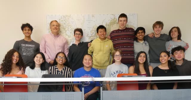Class photo of a group of students with their teacher in front of a large Arctic-centered world map in a classroom