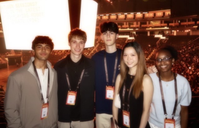 Evan and four other students wearing conference lanyards in the stands of a packed arena, a bright presentation screen and stage lighting behind them
