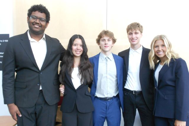 Evan, second from right, with four teammates in suits, smiling together against a pale wood wall