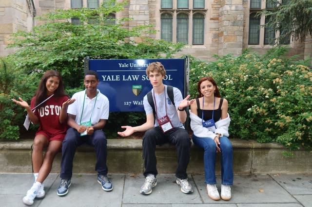 Evan Morris and three friends sitting on a ledge in front of the Yale Law School sign, shrugging for the camera