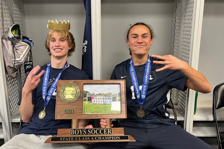 Evan Morris in a gold paper crown and a state championship medal, sitting beside a teammate with the Boys Soccer State Class A Champion plaque
